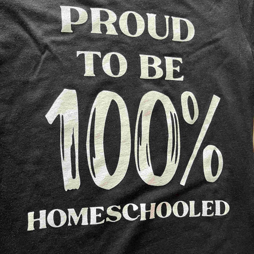 Close-up of a black tee with a white print reading 'Proud to be 100% homeschooled'.